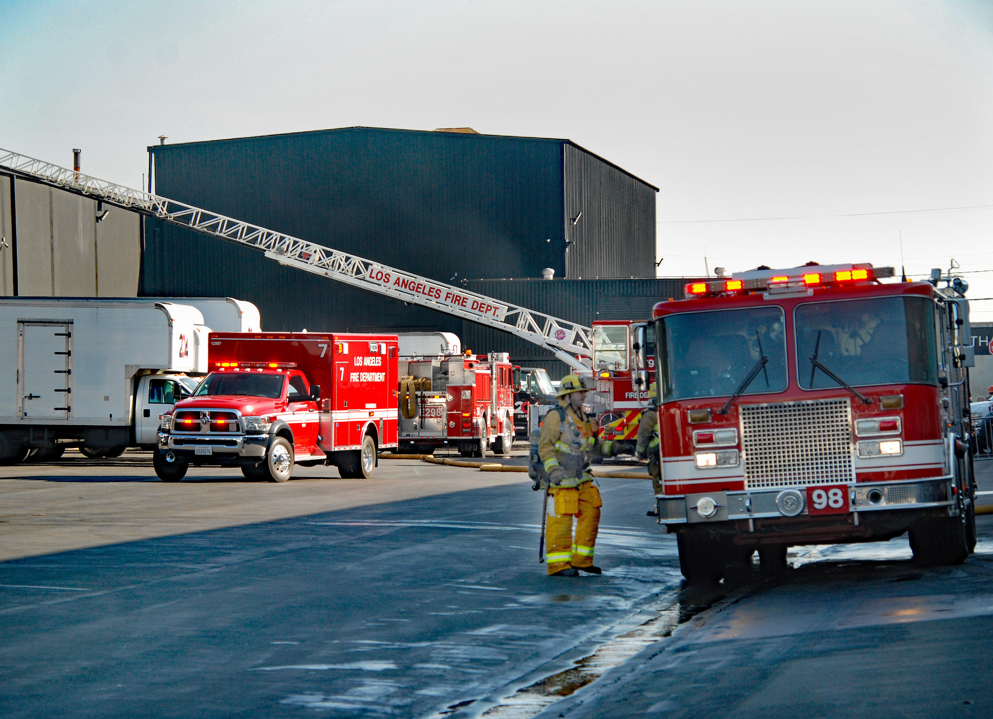 An LAFD ambulance, fire engine, and fire truck parked in front of an industrial building with the aerial ladder extending to the roof.