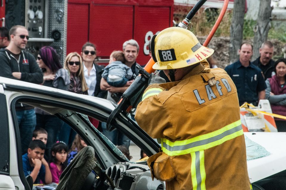 Firefighter performing a vehicle extrication demonstration with a crowd of members of the public watching in the background.