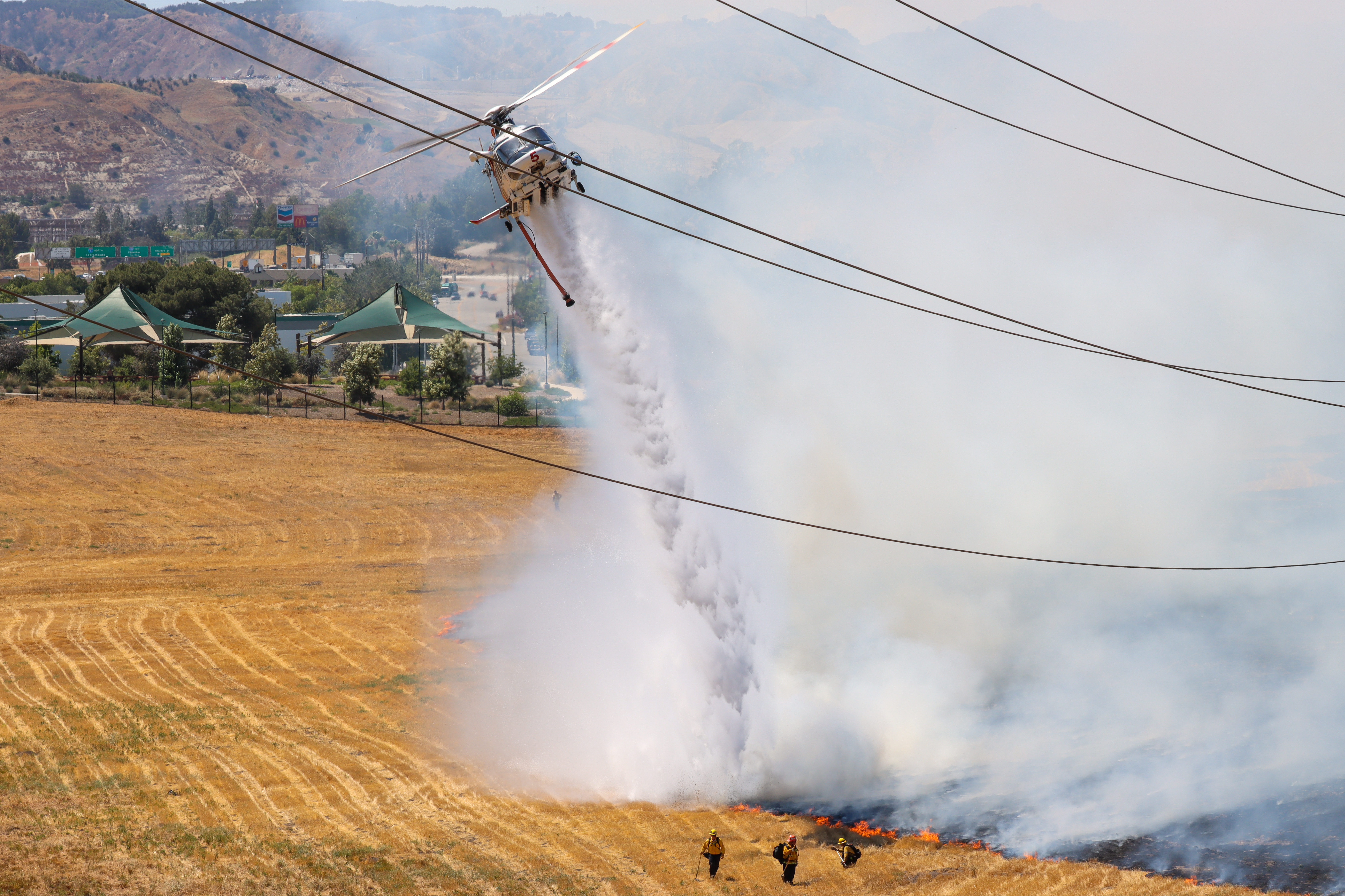 Helicopter dropping water on grass fire, with firefighters on the ground nearby