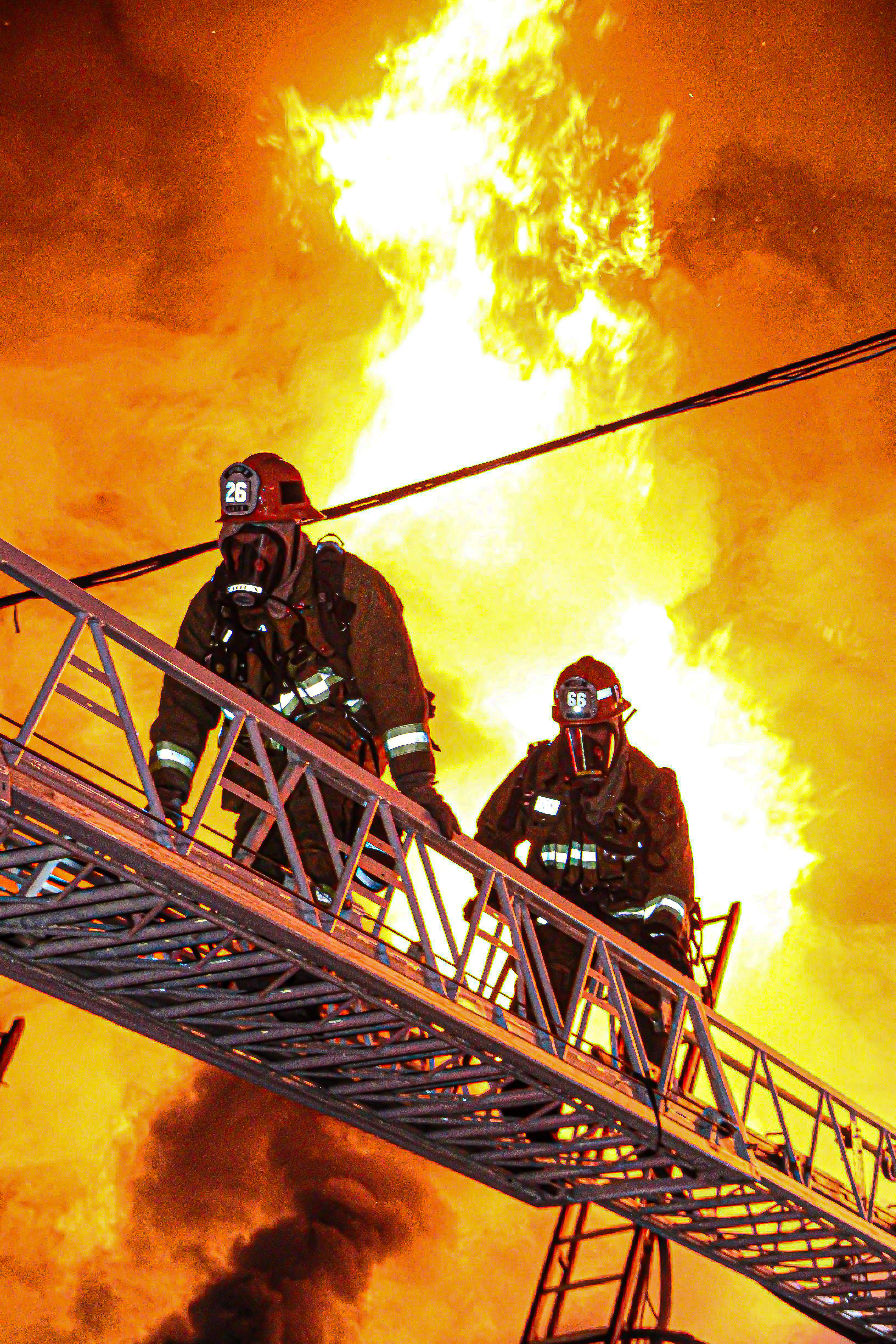 Two firefighters ascending aerial ladder with fire ranging in the back