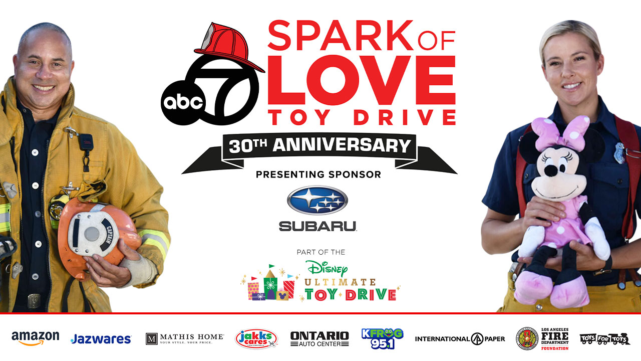 A smiling female firefighter holding a stuffed toy, standing alongside a smiling male firefighter holding a firefighting helmet and wearing protective gear.  Text: ABC 7 Spark of Love Toy Drive 30th Anniversary. Presenting sponsor Subaru. Part of the Disney Ultimate Toy Drive. Sponsor logos: Amazon, Jazwares, Mathis Home, Jakks, Ontario Auto Center, KFrog 95.1, International Paper, Los Angeles Fire Department Foundation, Toys For Tots.