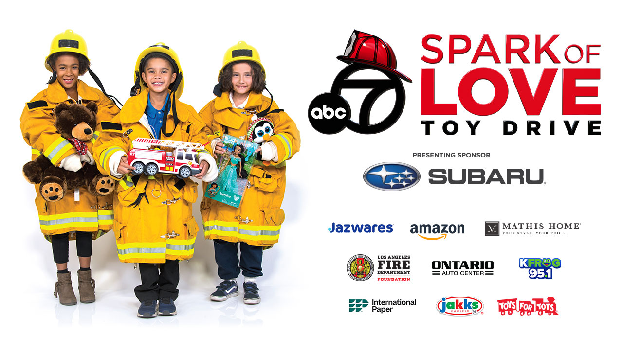 Three smiling children stand side by side, wearing firefighter helmets and coats and clutching toys. Logos for ABC7 Spark of Love Toy Drive, Subaru, Jazwares, Amazon, Mathis Home, Los Angeles Fire Department Foundation, Ontario Auto Center, KFROG 95.1, International Paper, Jakks, Toys For Tots.