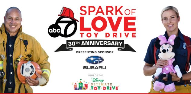A smiling female firefighter holding a stuffed toy, standing alongside a smiling male firefighter holding a firefighting helmet and wearing protective gear.  Text: ABC 7 Spark of Love Toy Drive 30th Anniversary. Presenting sponsor Subaru. Part of the Disney Ultimate Toy Drive. Sponsor logos: Amazon, Jazwares, Mathis Home, Jakks, Ontario Auto Center, KFrog 95.1, International Paper, Los Angeles Fire Department Foundation, Toys For Tots.